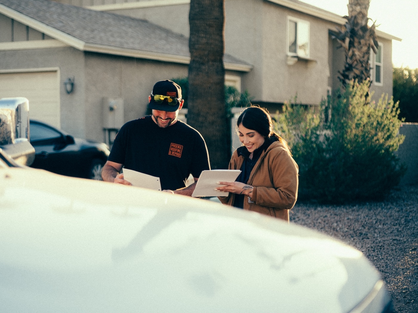 A contractor and a client smile while looking at paperwork outside near a house.