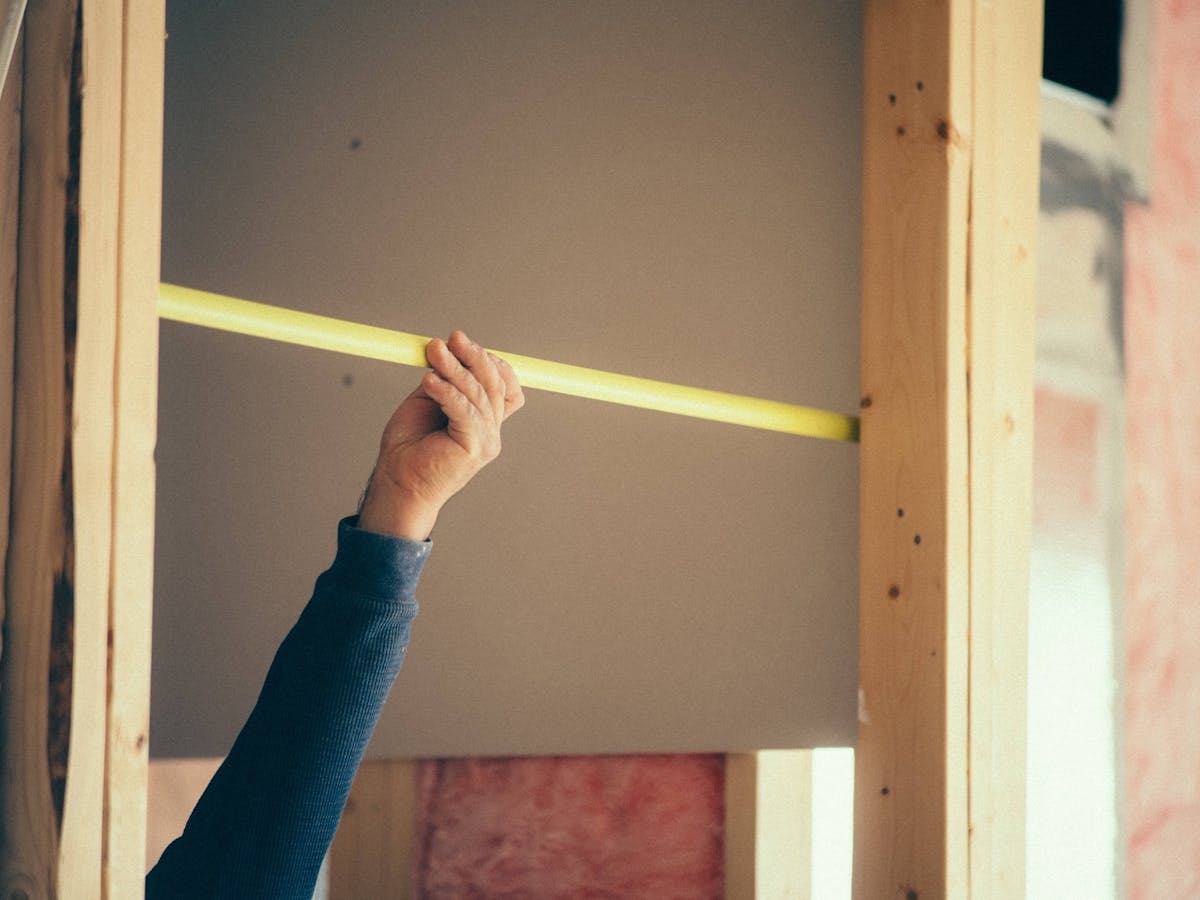 a person measures a section of drywall with a yellow measuring tape inside a wooden frame