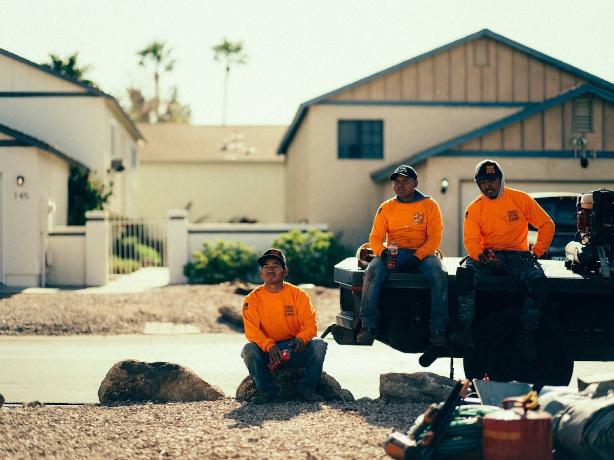 three construction workers wearing bright orange shirts take a break, with two sitting on a flatbed truck and one sitting on a rock in front of residential homes