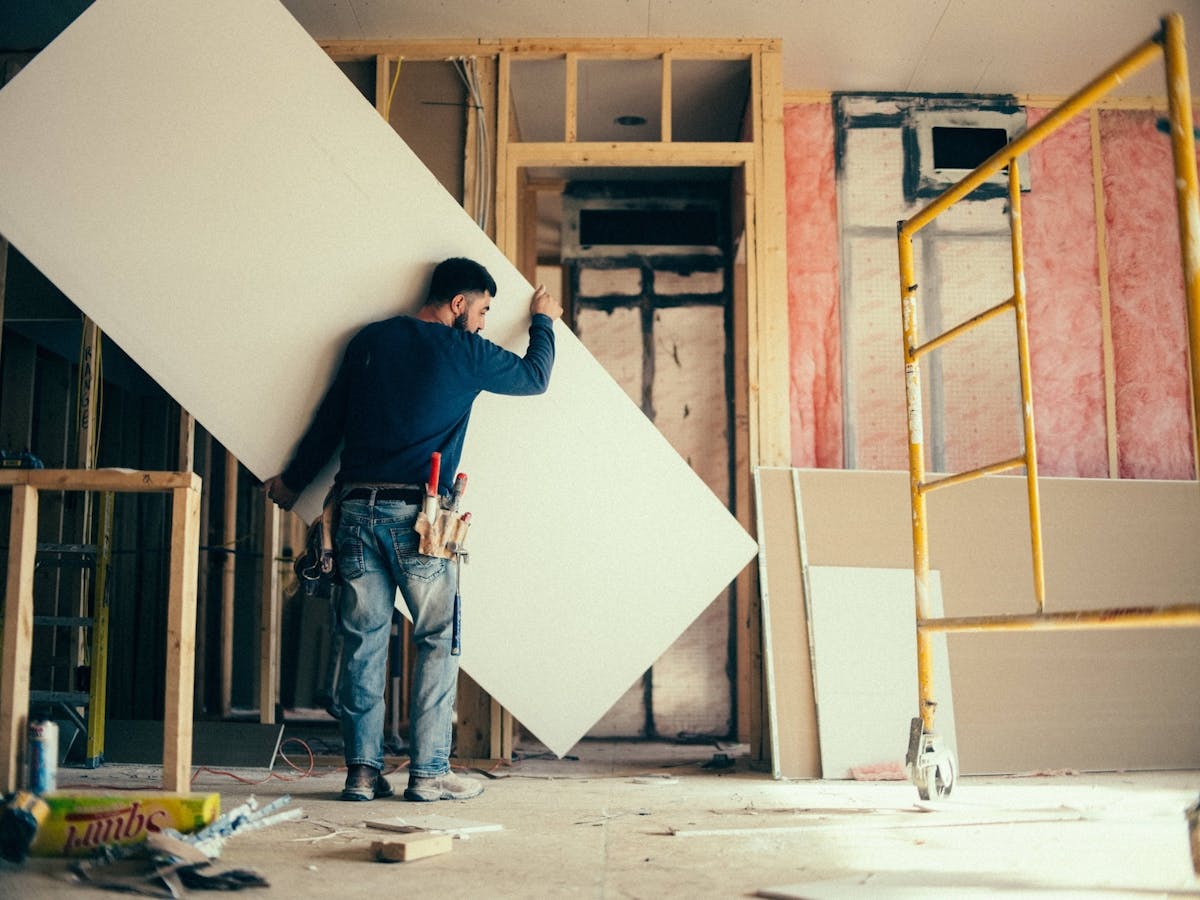 construction worker carries a large sheet of drywall inside a building under renovation, surrounded by exposed wooden framing, insulation, and tools