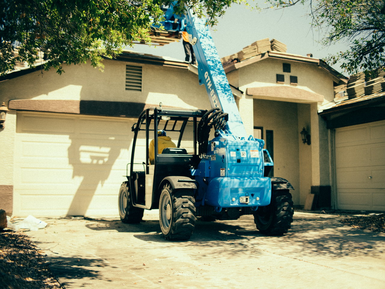 Blue telehandler lifting roofing materials to a residential rooftop during a roof replacement project