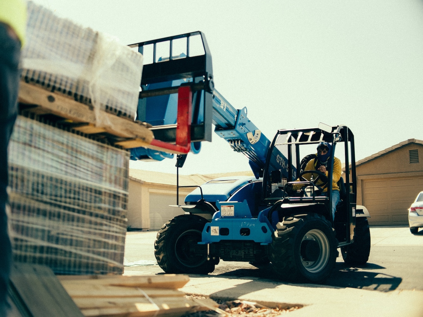 Equipment operator lifts palletized building materials with forklift on active residential construction site.