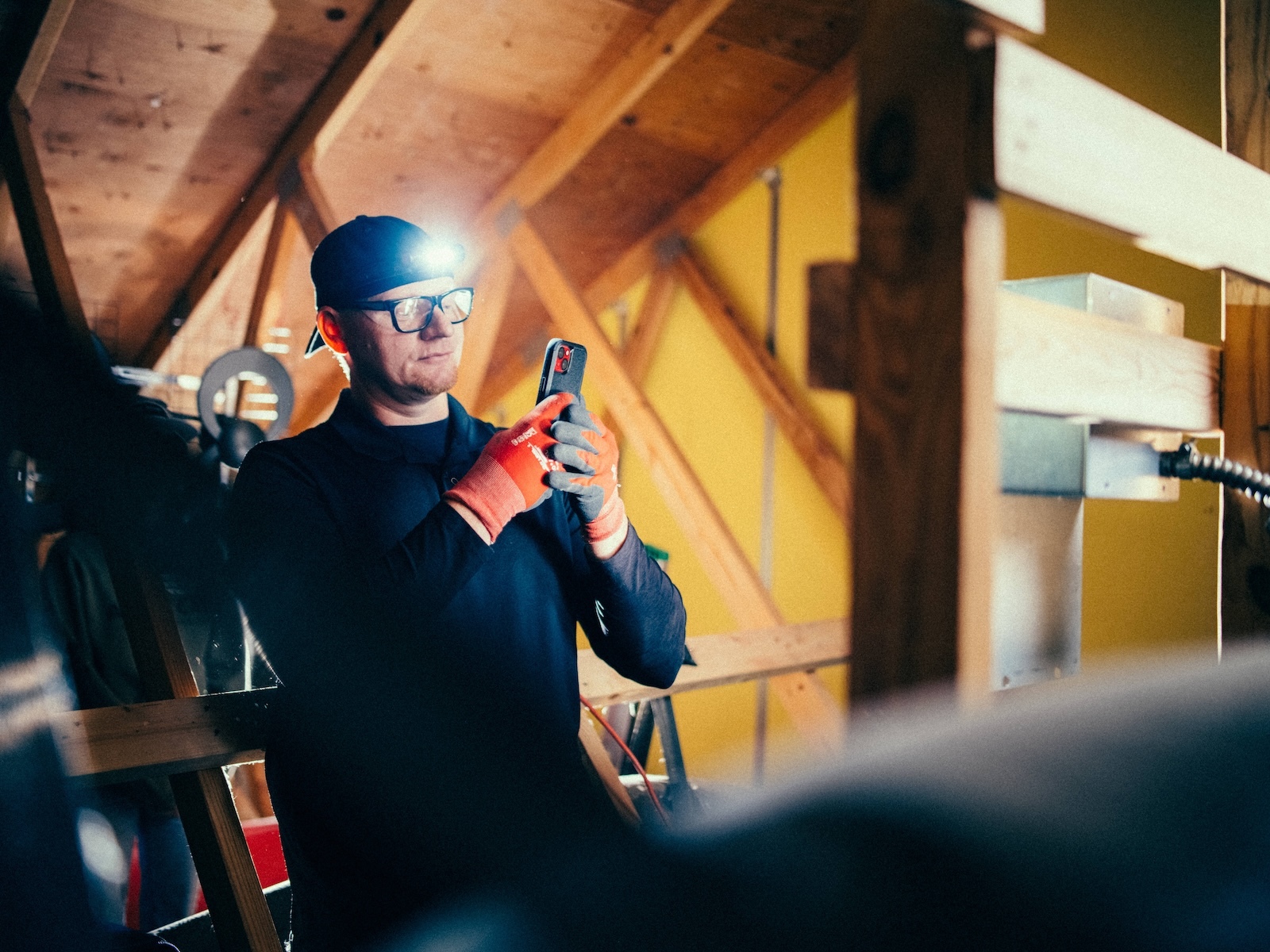 Technician documents attic electrical work with mobile app using headlamp for visibility.