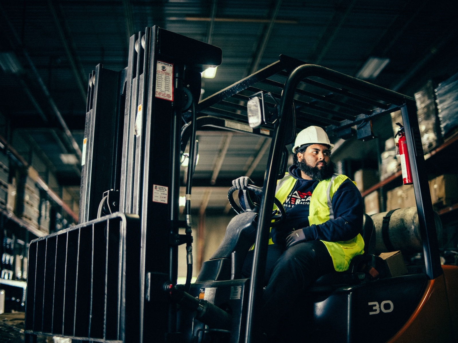 Warehouse worker operates forklift while moving materials through storage racks on industrial job site.