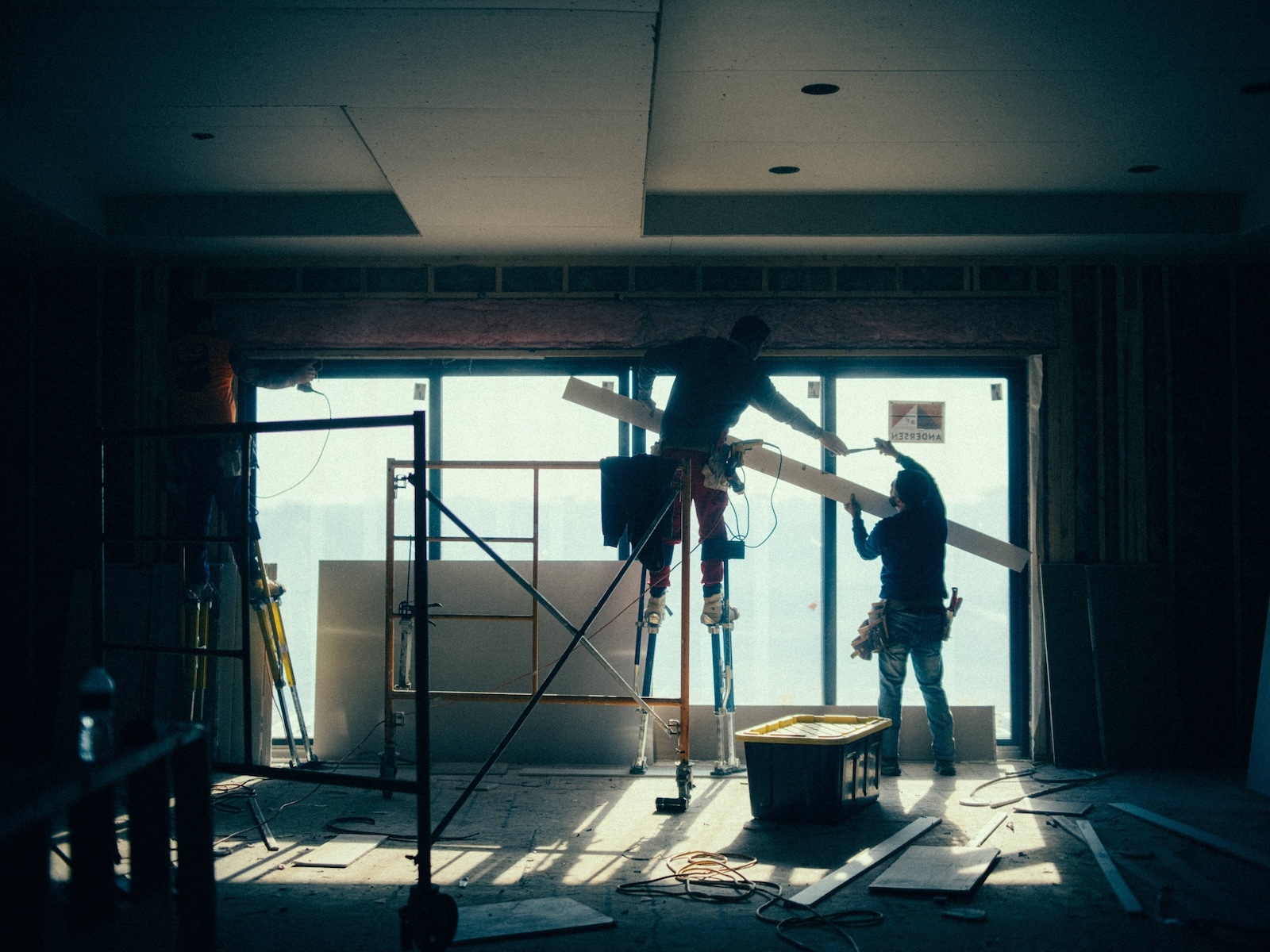 Framing crew installs ceiling material on stilts while another worker assists near large window opening.