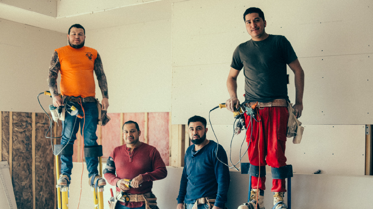 Four construction workers indoors, two standing on stilts with tools, preparing drywall installation.