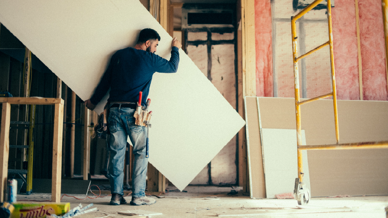 Construction worker carrying a large sheet of drywall inside a building under renovation.
